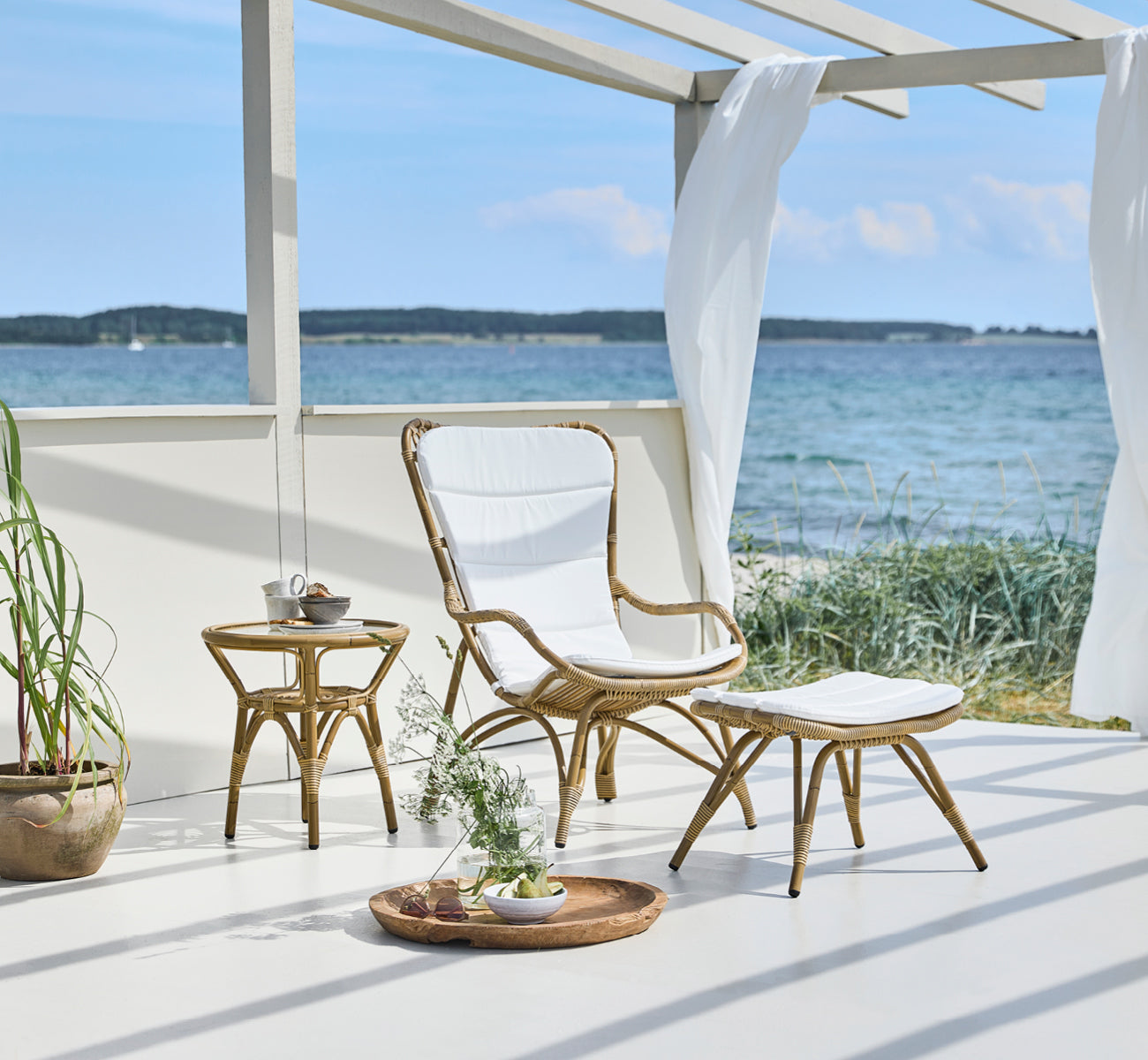 Esta es una imagen de estilo de vida de Monet Exterior Footstool i antique aluminium artfibre. En un pabellón junto al mar con sillón acolchado.