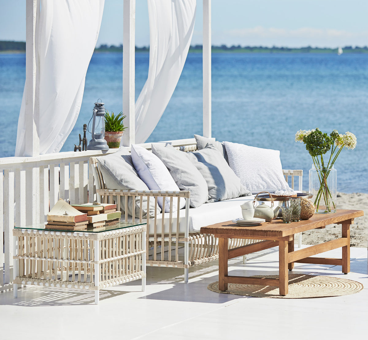 Esta es una imagen de estilo de vida de Caroline sofá exterior en dove White Aluminium ArtFibre, en una terraza de madera junto al agua con.