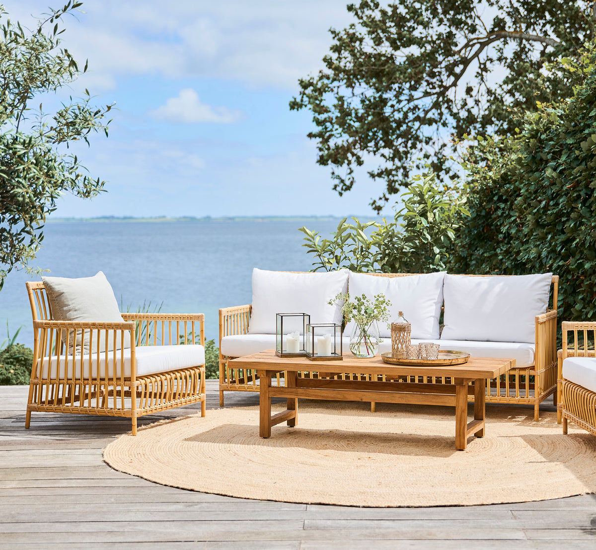 Esta es una imagen de estilo de vida de Caroline sofá exterior en natural Aluminium ArtFibre, en una terraza de madera junto al agua con vegetación.