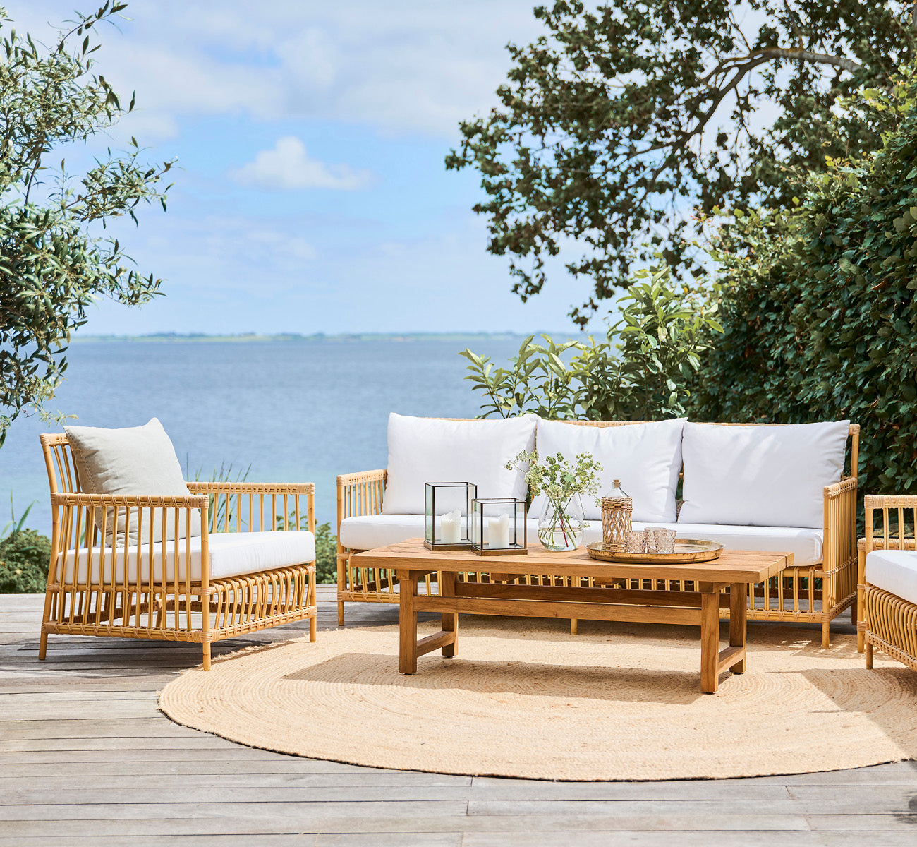 Esta es una imagen de estilo de vida de Caroline sofá exterior en natural Aluminium ArtFibre, en una terraza de madera junto al agua con vegetación.