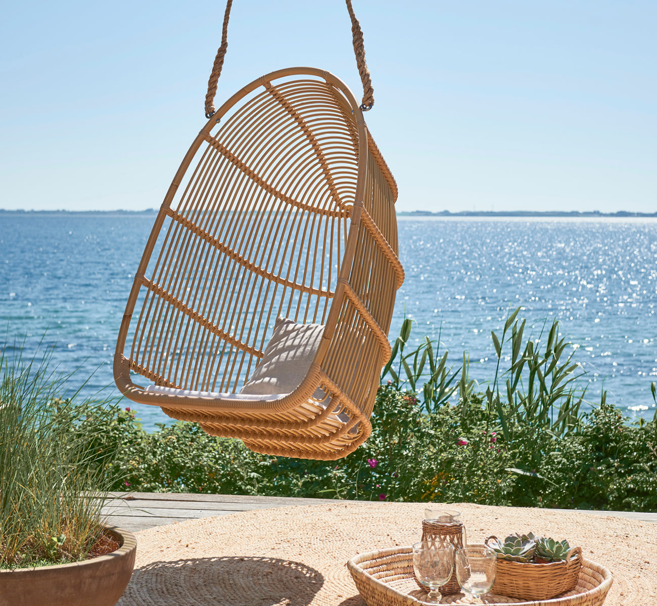 Esta es una imagen de estilo de vida de Renoir silla colgante exterior en natural aluminium artfibre. Colgando en una terraza junto al mar con bandeja y suculentas debajo.