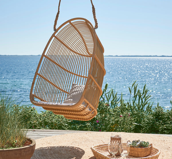 Esta es una imagen de estilo de vida de Renoir silla colgante exterior en natural aluminium artfibre. Colgando en una terraza junto al mar con bandeja y suculentas debajo.