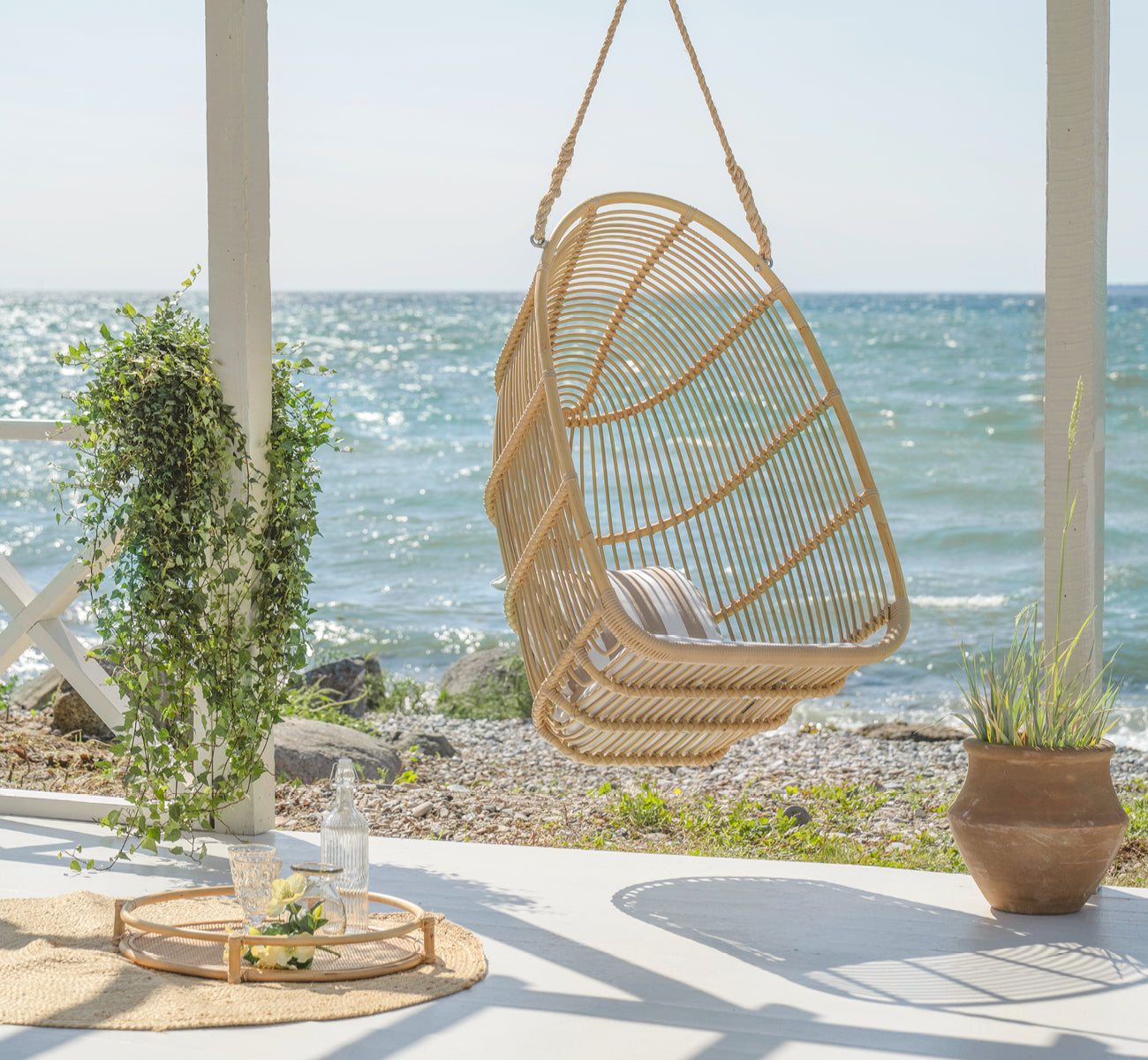 Esta es una imagen de estilo de vida de Renoir silla colgante exterior en natural aluminium artfibre. Colgando en una terraza junto al mar con bandeja y suculentas debajo.