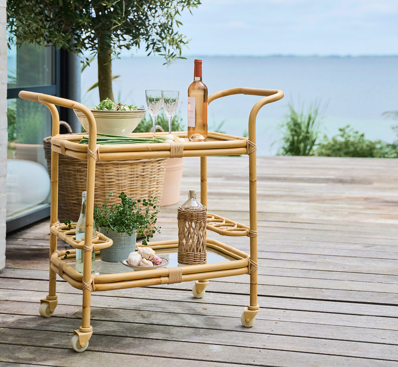 Esta es una imagen de estilo de vida de Carlo carrito exterior en natural Aluminium ArtFibre, en una terraza de madera junto al agua con vegetación.