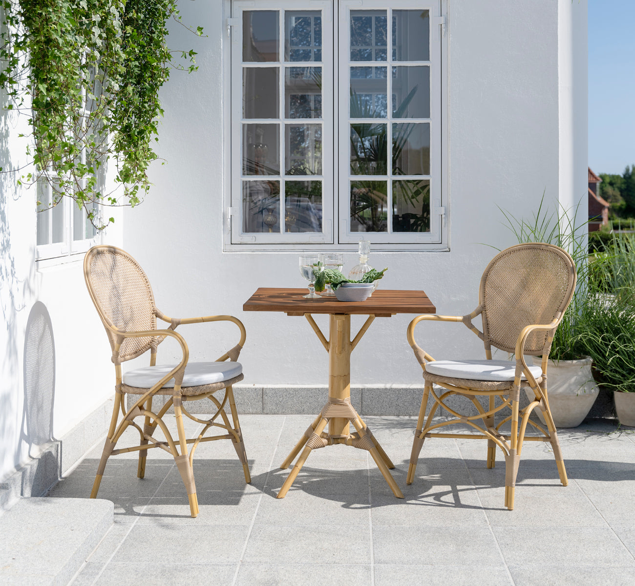 Esta es una imagen de estilo de vida de Nicole Cafe Table i natural aluminium artfibre. En un patio de piedra con sillas Ofelia y vegetación.