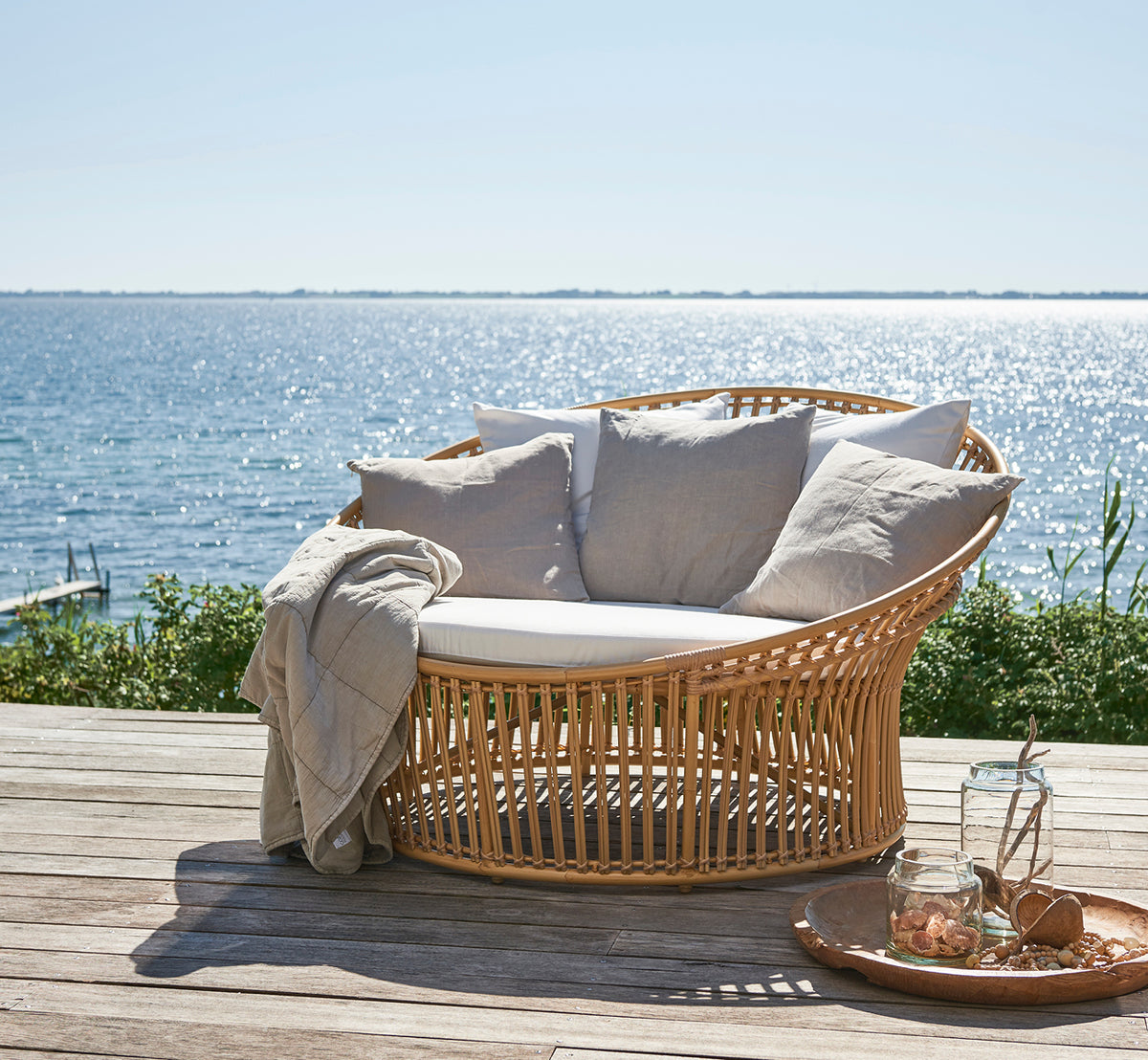 Esta es una imagen de estilo de vida de Olympia Nest Exterior i natural aluminium artfibre. En un pabellón de playa con cojines y vista al mar.