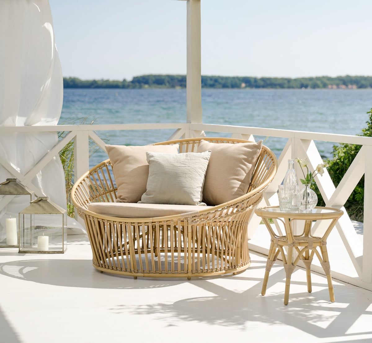 Esta es una imagen de estilo de vida de Olympia Nest Exterior i natural aluminium artfibre. En un pabellón junto al mar con cojines de lino.