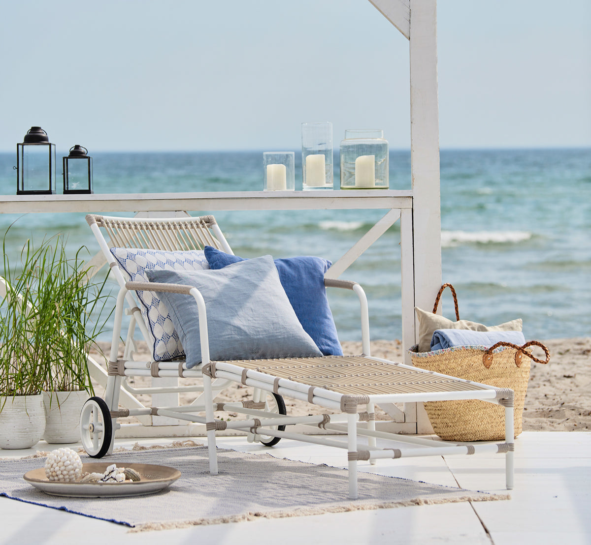 Esta es una imagen de estilo de vida de Josephine tumbona en dove White aluminium artfibre, en una cubierta de madera blanca junto a la playa bajo.