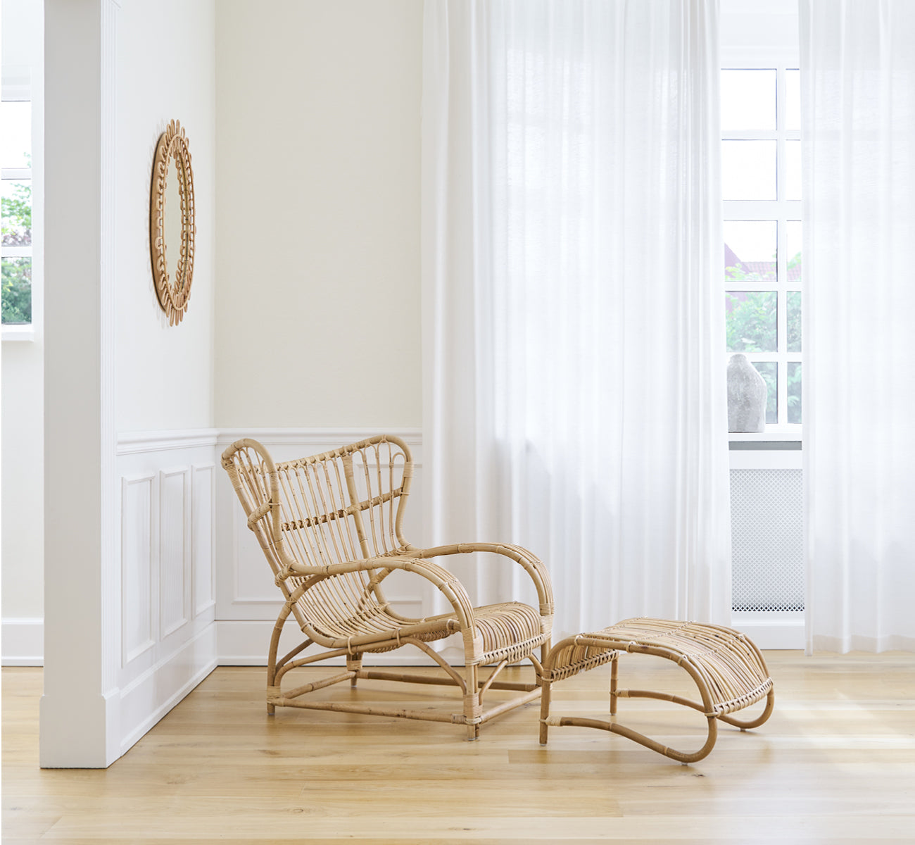 Esta es una imagen de estilo de vida de Teddy sillón lounge en polished natural rattan. Colocado sobre un suelo en espiga junto a una ventana con cortinas y un pequeño espejo.