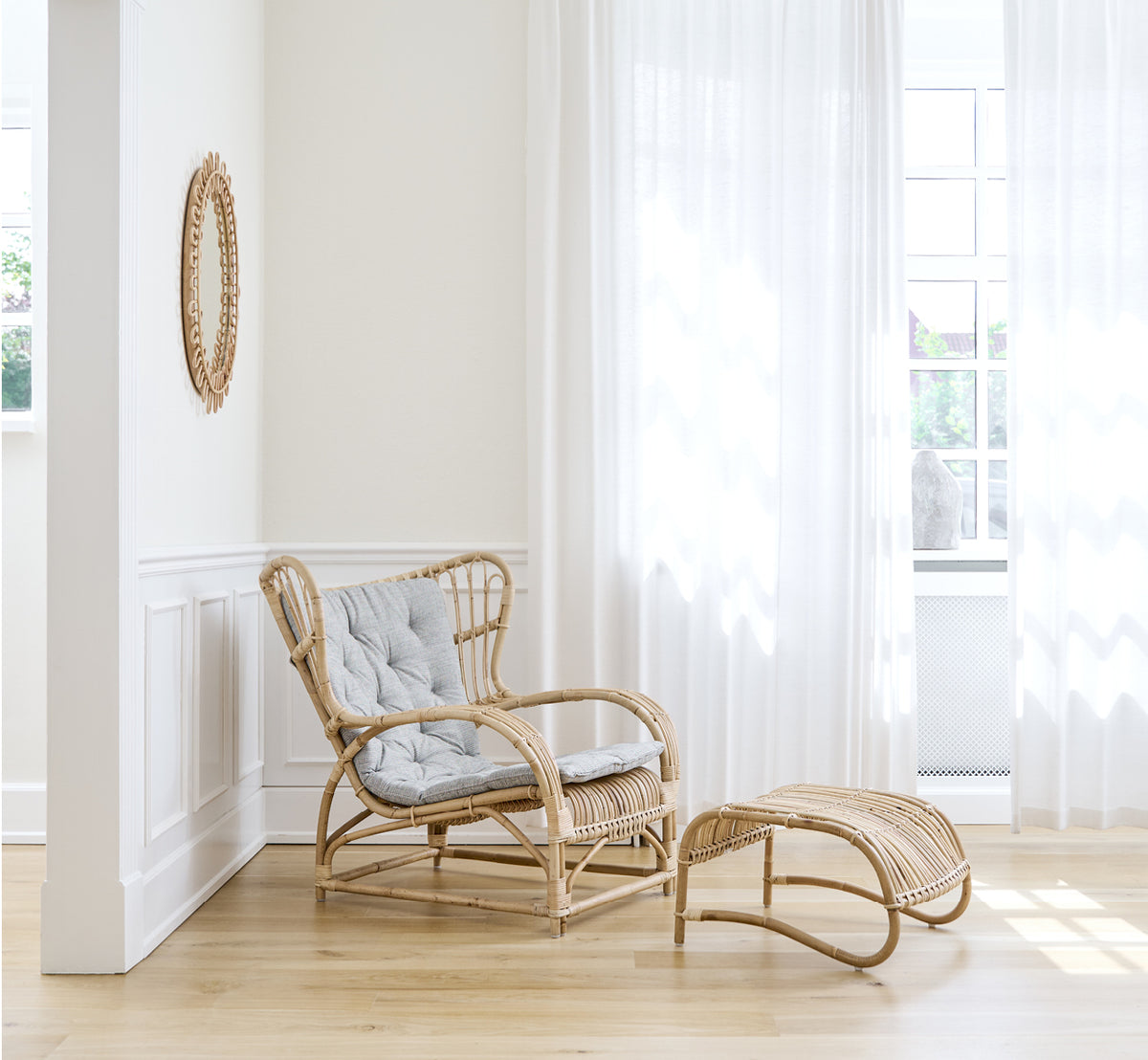 Esta es una imagen de estilo de vida de Teddy sillón lounge en polished natural rattan. Colocado sobre un suelo en espiga junto a una ventana con cortinas y un pequeño espejo.