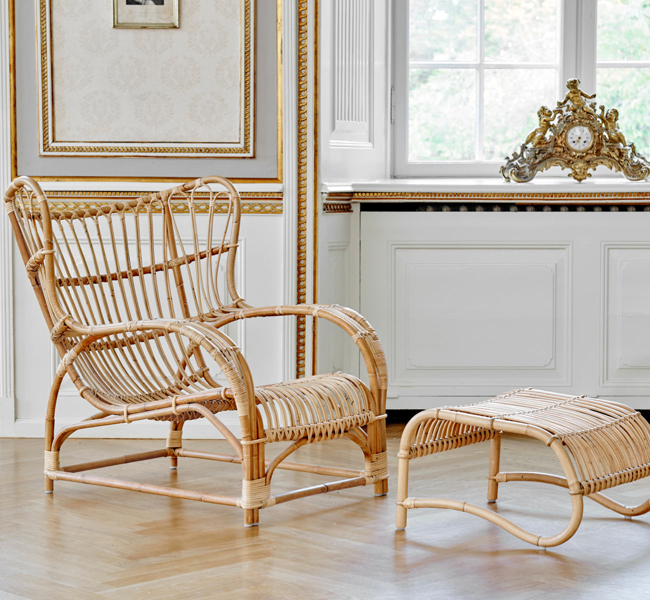 Esta es una imagen de estilo de vida de Teddy sillón lounge en polished natural rattan. Colocado sobre un suelo en espiga junto a una ventana con cortinas y un pequeño espejo.