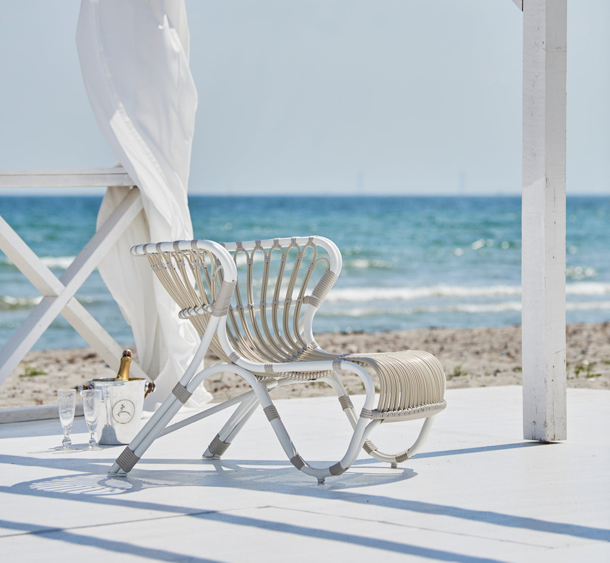 Esta es una imagen de estilo de vida de Fox sillón lounge exterior en dove white Aluminium ArtFibre, en una terraza de madera junto al agua con cañas.