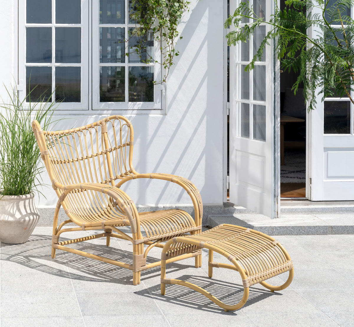 Esta es una imagen de estilo de vida de Teddy sillón lounge exterior en natural aluminium artfibre. Colocado en una terraza pavimentada soleada junto a un umbral con marco blanco.