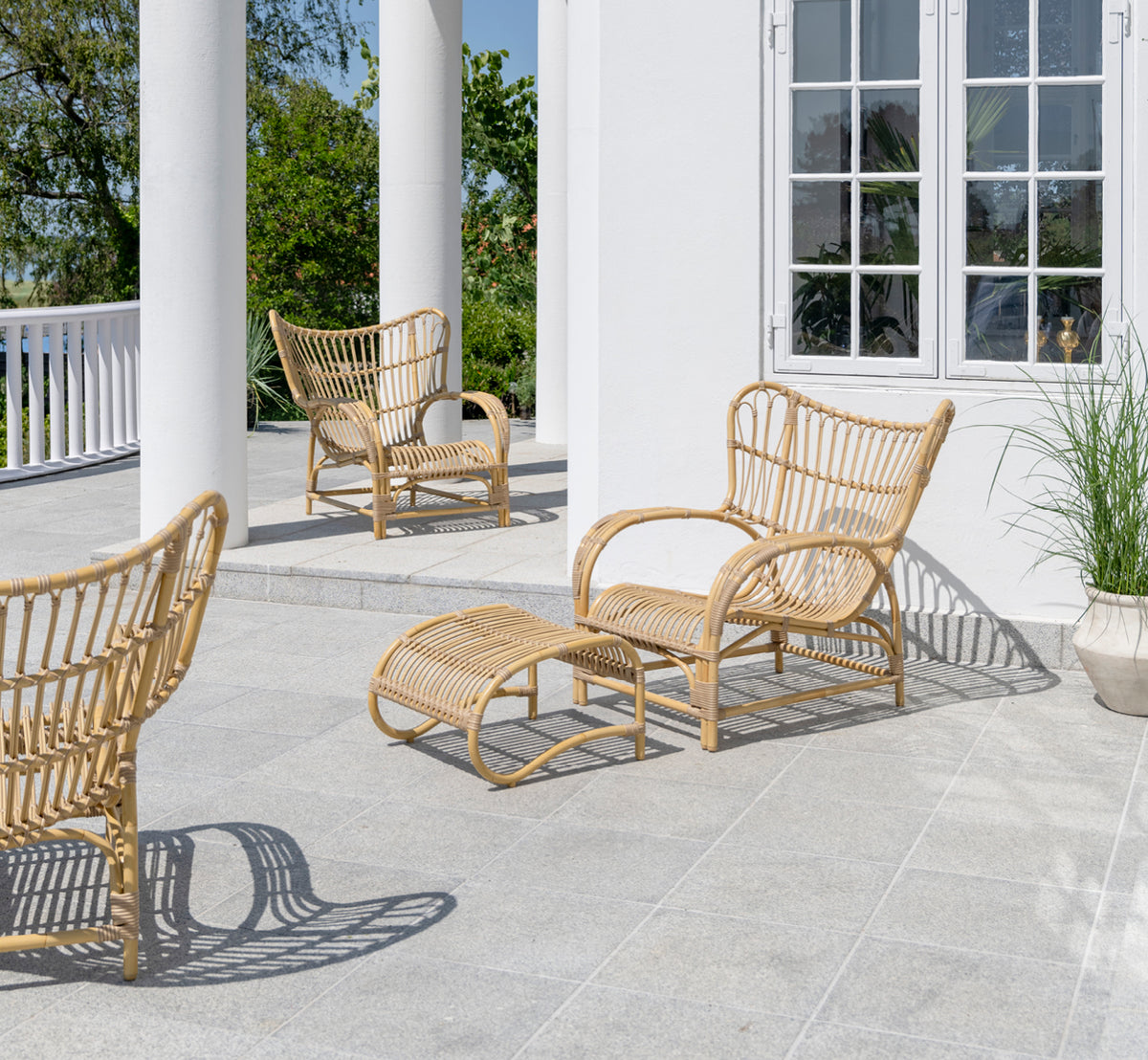 Esta es una imagen de estilo de vida de Teddy sillón lounge exterior en natural aluminium artfibre. Colocado en una terraza pavimentada soleada junto a un umbral con marco blanco.