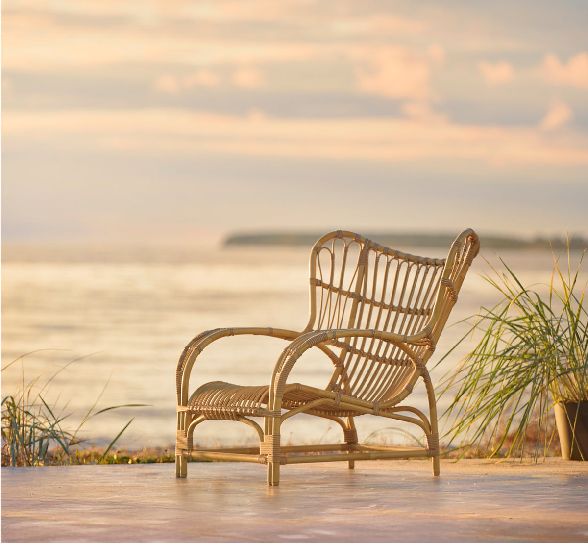 Esta es una imagen de estilo de vida de Teddy sillón lounge exterior en natural aluminium artfibre. Colocado en una terraza pavimentada soleada junto a un umbral con marco blanco.