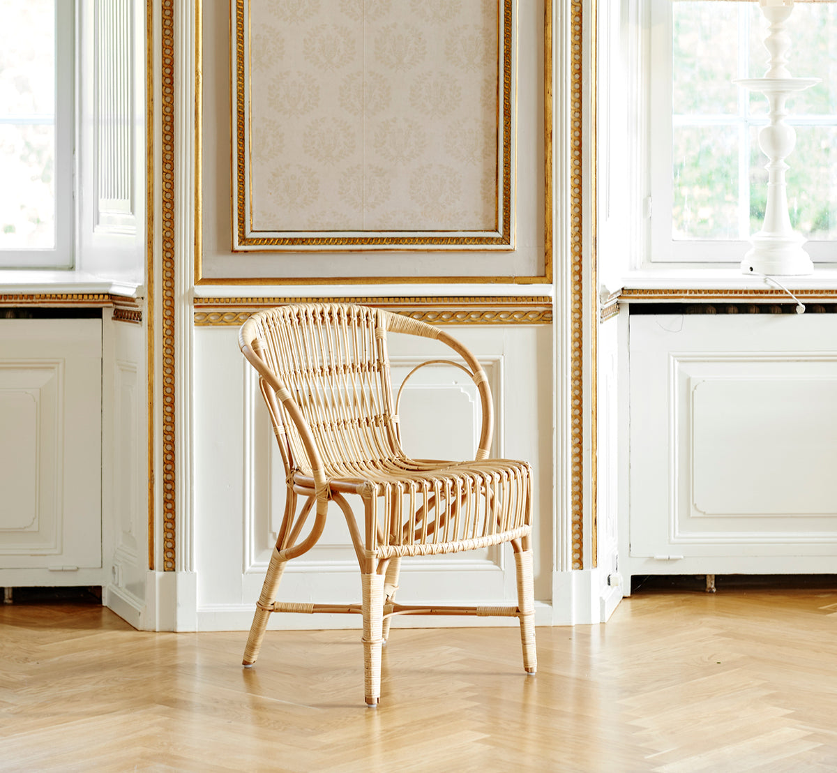 Esta es una imagen de estilo de vida de Robert silla de comedor en polished natural rattan. Colocada junto a una mesa de pedestal con tulipanes sobre una alfombra de yute.