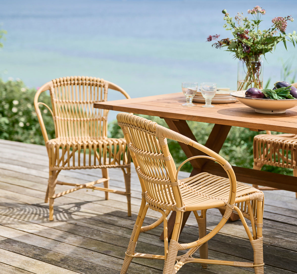 Esta es una imagen de estilo de vida de Robert silla de comedor exterior en natural aluminium artfibre. Dispuestas alrededor de una mesa de teca en una cubierta de madera con vistas al mar.