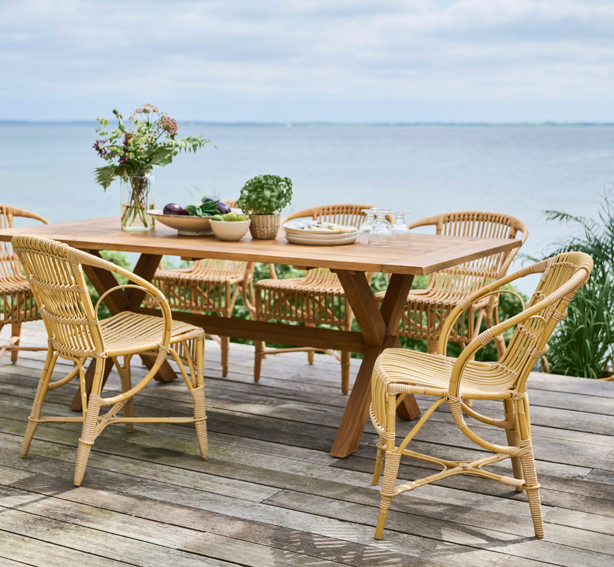 Esta es una imagen de estilo de vida de Robert silla de comedor exterior en natural aluminium artfibre. Dispuestas alrededor de una mesa de teca en una cubierta de madera con vistas al mar.