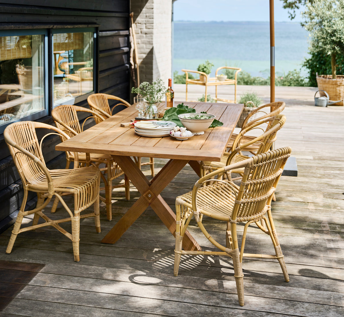 Esta es una imagen de estilo de vida de Robert silla de comedor exterior en natural aluminium artfibre. Dispuestas alrededor de una mesa de teca en una cubierta de madera con vistas al mar.