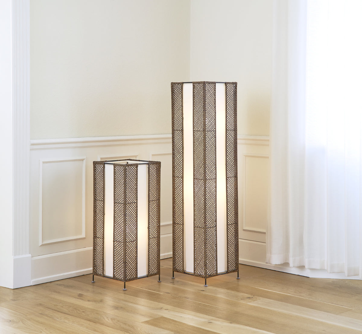 Esta es una imagen de estilo de vida de Yamakawa Lampshade | 75 cm en hazelnut rattan iron frame. Mostrada junto a una lámpara compañera alta en un rincón luminoso con paneles.